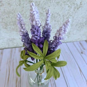 May include: A clear glass vase holds a bouquet of felt flowers. The flowers are a gradient of purple and lavender, with green felt leaves. The vase sits on a white wooden surface.