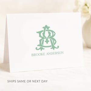 May include: A white folded note card featuring a sage green monogram of the letters B and A in a decorative serif font, with the name BROOKE ANDERSON printed in sage green capital letters directly underneath the monogram design.
