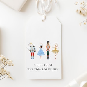 May include: White gift tag with a ribbon, featuring illustrations of a mouse, ballerina, nutcracker, and fairy. The tag reads "A GIFT FROM THE EDWARDS FAMILY". The tag is surrounded by wrapped gifts and floral accents.