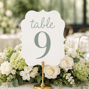 May include: A white table number sign with a scalloped edge featuring the word table and the number 9 in sage green text. The sign is mounted on a gold metal stand and is positioned in the center of a floral arrangement of white roses.