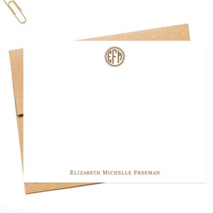 May include: A white rectangular stationery card with a brown circular monogram featuring the letters EFM at the top center and the name ELIZABETH MICHELLE FREEMAN printed in brown at the bottom, resting on a brown envelope with two paper clips.