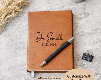 May include: A brown leather journal with the name "Dr. Smith" and "Ph.D. 2026" printed on the cover. A black pen rests on the journal. A small dish with gold rings and a binder clip are also visible.