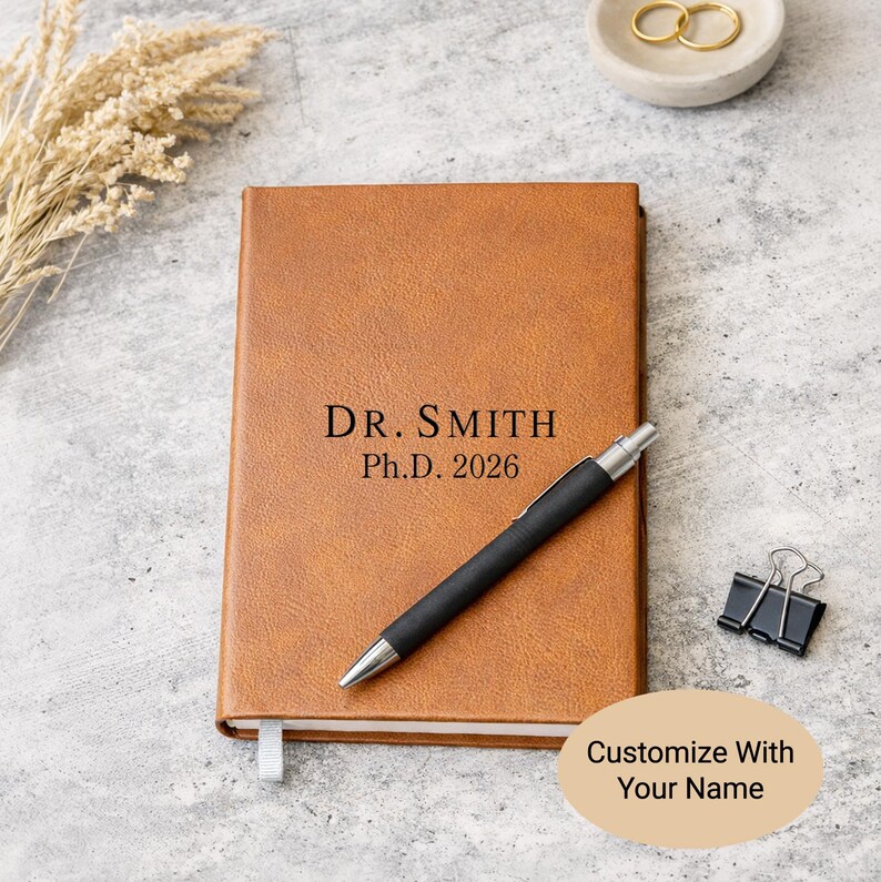 May include: A tan notebook with a textured cover featuring the black engraved text DR. SMITH Ph.D. 2026. A black and silver ballpoint pen rests diagonally across the front cover. A black metal binder clip is positioned to the right of the notebook.