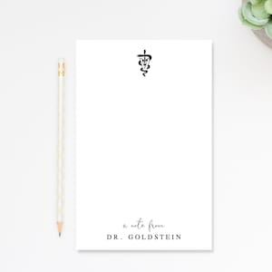 May include: A white rectangular notepad featuring a black veterinary caduceus symbol at the top and the text "a note from DR. GOLDSTEIN" at the bottom, accompanied by a light-colored wooden pencil positioned vertically along the left side.