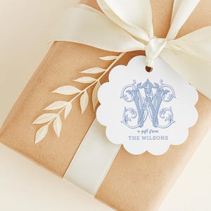 May include: A brown paper gift box wrapped with a cream-colored ribbon and a white scalloped gift tag. The tag features a blue ornate monogram letter W and the text a gift from THE WILSONS, accented by a small white leaf sprig.