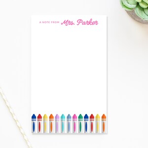 May include: White notepad with the text "A NOTE FROM Mrs. Parker" in pink. The bottom of the notepad features a colorful crayon border. The notepad is rectangular and has a clean, minimalist design.