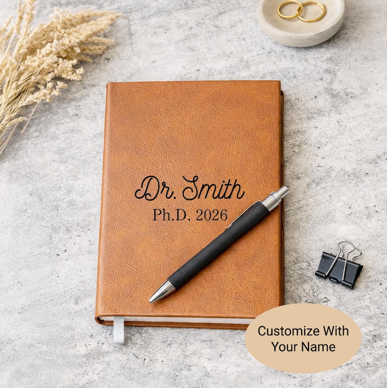 May include: A tan leather-textured notebook featuring the black engraved text Dr. Smith Ph.D. 2026 on the front cover. A black ballpoint pen with a silver tip and clip rests diagonally across the notebook. A black metal binder clip is nearby.