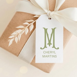 May include: A brown paper gift box wrapped with a cream-colored satin ribbon and a white rectangular gift tag. The tag features a large sage green letter M and the text CHERYL MARTINS in sage green. A dried white leaf sprig is tucked under the ribbon.