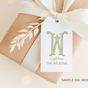 May include: A rectangular gift tag with a white background featuring a large green letter W, the text a gift from THE WILSONS, and a sprig of white leaves. The tag is tied to a brown paper wrapped gift with a wide white satin ribbon bow.