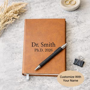 May include: A tan leather-like notebook with the black text Dr. Smith Ph.D. 2026 printed on the cover. A black ballpoint pen with a silver clip and tip rests diagonally across the notebook. A black metal binder clip is positioned to the right.