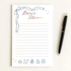 May include: A white notepad with blue and red nautical-themed illustrations and the text "Becca's Notes". The notepad has horizontal lines for writing and a blue ribbon design. A black pen with gold accents rests on the right.