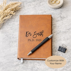 May include: A tan leather-textured notebook featuring the black printed text Dr. Smith Ph.D. 2026. A black ballpoint pen rests diagonally across the cover. A small black metal binder clip and a white ribbon bookmark are attached to the notebook.