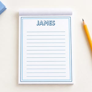 May include: A white notepad with a blue double-line border and the name JAMES printed in blue at the top. The notepad features horizontal blue lines for writing. A yellow wooden pencil with a pink eraser is positioned to the right of the pad.