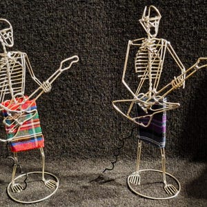 May include: Two wire skeleton figures playing guitars. One wears a red, green, and blue tartan kilt, the other a purple, black, and blue tartan kilt. The guitars are also made of wire, with a cable attached to one.