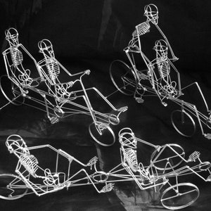 May include: Four wire sculptures of skeletons riding recumbent tricycles. The skeletons are made of silver wire and are posed in a variety of riding positions. The tricycles are also made of silver wire and have three wheels.