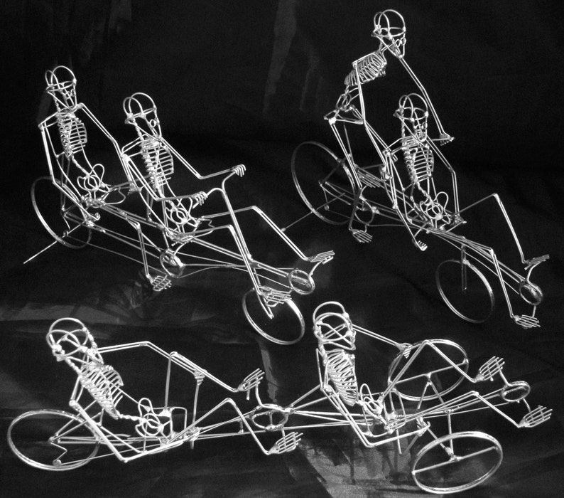May include: Four wire sculptures of skeletons riding recumbent tricycles. The skeletons are made of silver wire and are posed in a variety of riding positions. The tricycles are also made of silver wire and have three wheels.