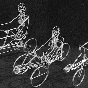 May include: Three wire sculptures of skeletons riding tricycles. The skeletons are made of thin wire and are posed in a riding position. The tricycles are also made of wire and have three wheels.