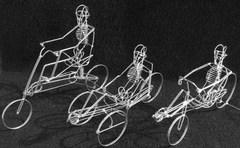 May include: Three wire sculptures of skeletons riding tricycles. The skeletons are made of thin wire and are posed in a riding position. The tricycles are also made of wire and have three wheels.
