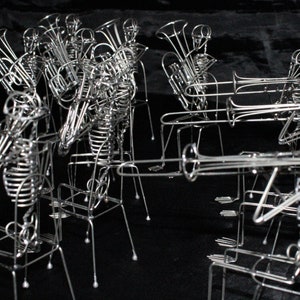 May include: A group of wire sculptures of skeletons playing musical instruments. The skeletons are posed as if they are playing a brass band. The sculptures are made of silver wire and are arranged in a row.