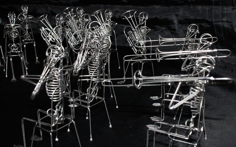 May include: A group of wire sculptures of skeletons playing musical instruments. The skeletons are posed as if they are playing a brass band. The sculptures are made of silver wire and are arranged in a row.