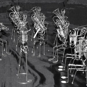 May include: A group of silver skeleton figures playing musical instruments. The figures are made of wire and are posed in a band formation. The instruments include a trumpet, a trombone, a tuba, a clarinet, and a saxophone.