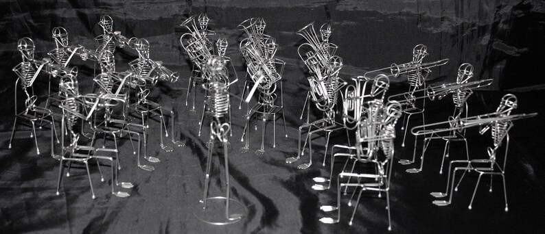 May include: A group of silver skeleton figures playing musical instruments. The figures are made of wire and are posed in a band formation. The instruments include a trumpet, a trombone, a tuba, a clarinet, and a saxophone.