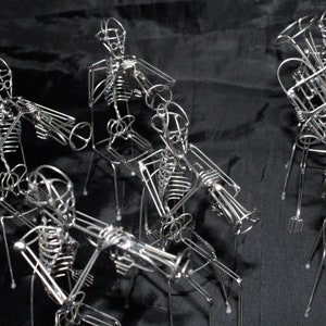 May include: A group of wire sculptures of skeletons playing musical instruments. The skeletons are posed in a band formation, each holding a different instrument, such as a trumpet, trombone, and tuba. The sculptures are made of thin wire and are very detailed.