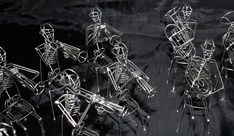 May include: A group of wire sculptures of skeletons playing musical instruments. The skeletons are posed in a band formation, each holding a different instrument, such as a trumpet, trombone, and tuba. The sculptures are made of thin wire and are very detailed.