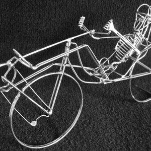 May include: A silver wire sculpture of a recumbent bicycle with a skeletal rider. The bicycle has a large front wheel and a smaller rear wheel.