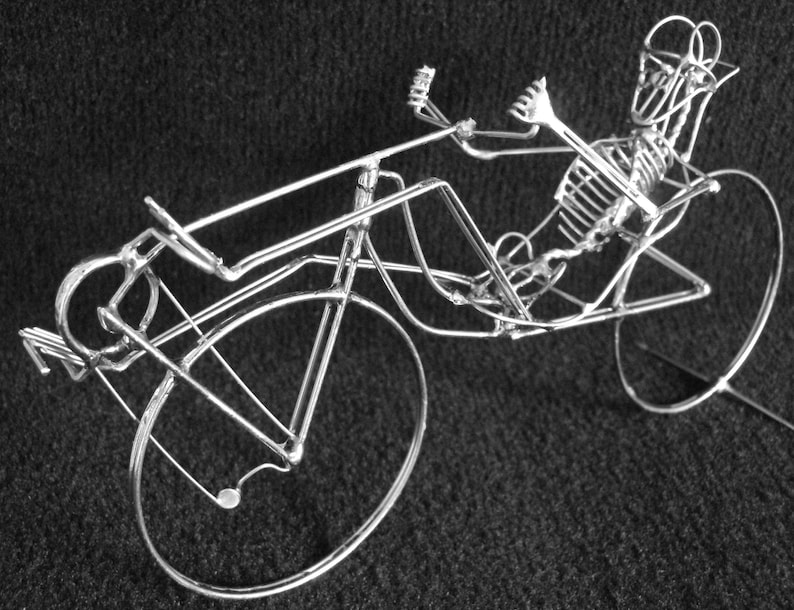 May include: A silver wire sculpture of a recumbent bicycle with a skeletal rider. The bicycle has a large front wheel and a smaller rear wheel.