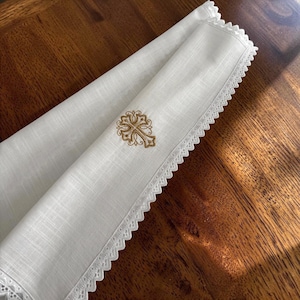 May include: A white linen cloth featuring a decorative scalloped lace border along the edge. The fabric is embroidered with a gold-colored cross design centered on the folded material, showcasing intricate stitching and a clean, textured finish.