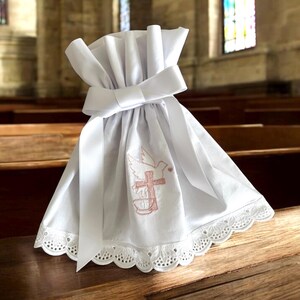 May include: White fabric bag with a white satin ribbon and a pink embroidered dove and cross design. The bag is sitting on a wooden surface.