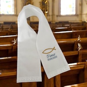 Peut inclure: Une étole de baptême blanche ornée d'un symbole de poisson doré brodé, du prénom Timo et de la date 31.08.2025 en fil bleu. L'étole est terminée par une délicate bordure en dentelle blanche sur le bas des deux pans suspendus.