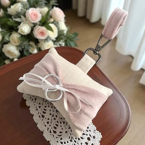 May include: A square ring pillow with a light pink accent and a matching handle. Two silver rings are tied with a white ribbon. The pillow is on a wooden surface, with a lace doily and a bouquet of roses in the background.