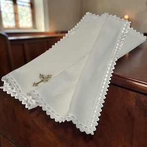 Könnte beinhalten: Ein weißes Leinentuch mit einer dekorativen, gebogten Spitzenborte und einem kleinen, goldfarben bestickten Kreuz. Der Stoff ist gefaltet, um die filigrane Handarbeit und die detaillierten Nähte entlang der Textilkanten zu zeigen.