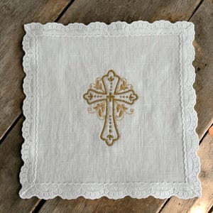 May include: A white linen handkerchief with a gold embroidered cross at its centre. The edges are trimmed with delicate, scalloped lace. The cross design features intricate detailing, creating an elegant aesthetic.