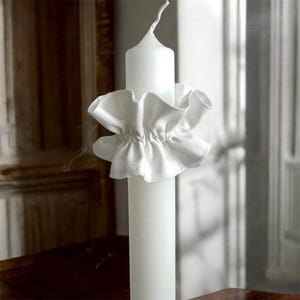 May include: A white pillar candle with a ruffled fabric candle ring. The candle is cylindrical and has a slightly curved wick. The fabric ring is gathered and adds a decorative element to the candle.