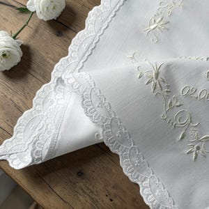 Peut inclure: Un chemin de table blanc orné de broderies florales couleur crème et d'une bordure en dentelle festonnée. La broderie présente des motifs de feuilles délicats et le texte 02.06, mettant en valeur un travail d'aiguille détaillé sur un tissu lisse.
