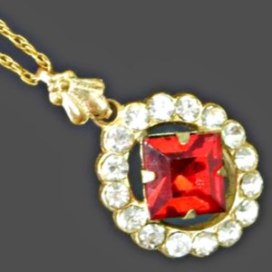 May include: A gold-toned necklace featuring a square, red gemstone pendant surrounded by small, clear crystals. The pendant hangs from a delicate gold chain. The necklace has a vintage or antique aesthetic.