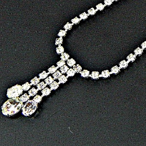 May include: A silver-toned necklace with a cascading design. The necklace features multiple strands of small, round, clear crystals, culminating in three larger, oval-shaped crystals at the bottom. The jewelry is set against a dark background.