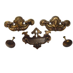 May include: A collection of antique brass drawer pulls and knobs. The set comprises two ornate pulls with a floral design, a central pull with a keyhole, and two round knobs. The hardware has a vintage, aged appearance.
