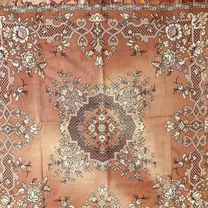 Puede incluir: Un mantel cuadrado, de color marrón, bordado con un patrón floral y un medallón central. El mantel tiene un borde de flecos.