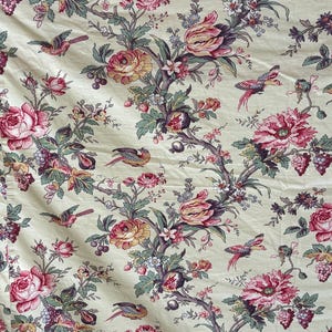 Op de afbeelding: Een lichtcrème stof met een herhalend patroon van roze en gele rozen, tulpen en kleine witte bloemen, verweven met donkergroene bladeren, paarse druiven en kleine kleurrijke vogels die tussen de takken door vliegen.