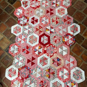 Pode incluir: Uma colcha feita à mão com um padrão hexagonal repetitivo composto por vários retalhos de tecido vermelho e branco. O design incorpora estampas florais, xadrez e vermelho sólido dispostas num mosaico geométrico, criando um têxtil vibrante.