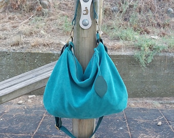 Op de afbeelding: Een tealblauwe suède handtas met een leren label waarop "Leather Tribe" staat. De tas heeft een lange band en hangt aan een houten paal.