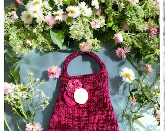 May include: A knitted magenta handbag with a circular handle and a decorative flower. The bag is surrounded by white and pink daisies and other greenery, set against a light blue background. The bag's texture is visible.