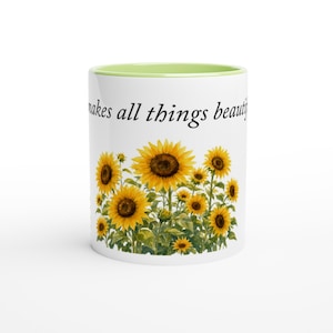 Puede incluir: Una taza de cerámica blanca con interior y borde de color verde claro. El exterior presenta una ilustración estilo acuarela de girasoles amarillos con tallos y hojas verdes. El texto "makes all things beautiful" está impreso en negro.
