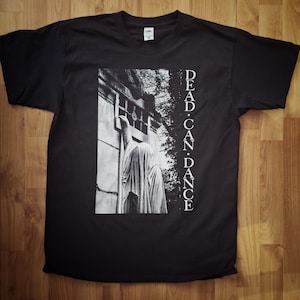 Può includere: Maglietta nera con una grafica bianca di una statua di fronte a un edificio. Il testo "DEAD. CAN. DANCE" è stampato verticalmente sul lato destro della grafica.