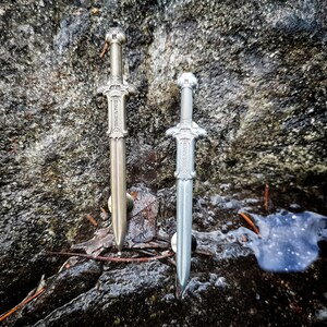Pu&ograve; includere: Due spade color argento con disegni intricati su uno sfondo roccioso. Le spade sono lunghe circa 25 cm e hanno una punta appuntita.