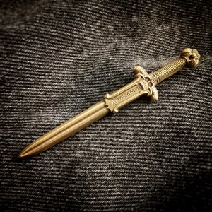 May include: A gold-coloured sword with intricate details and a decorative handle. The sword is lying on a dark grey textured surface.